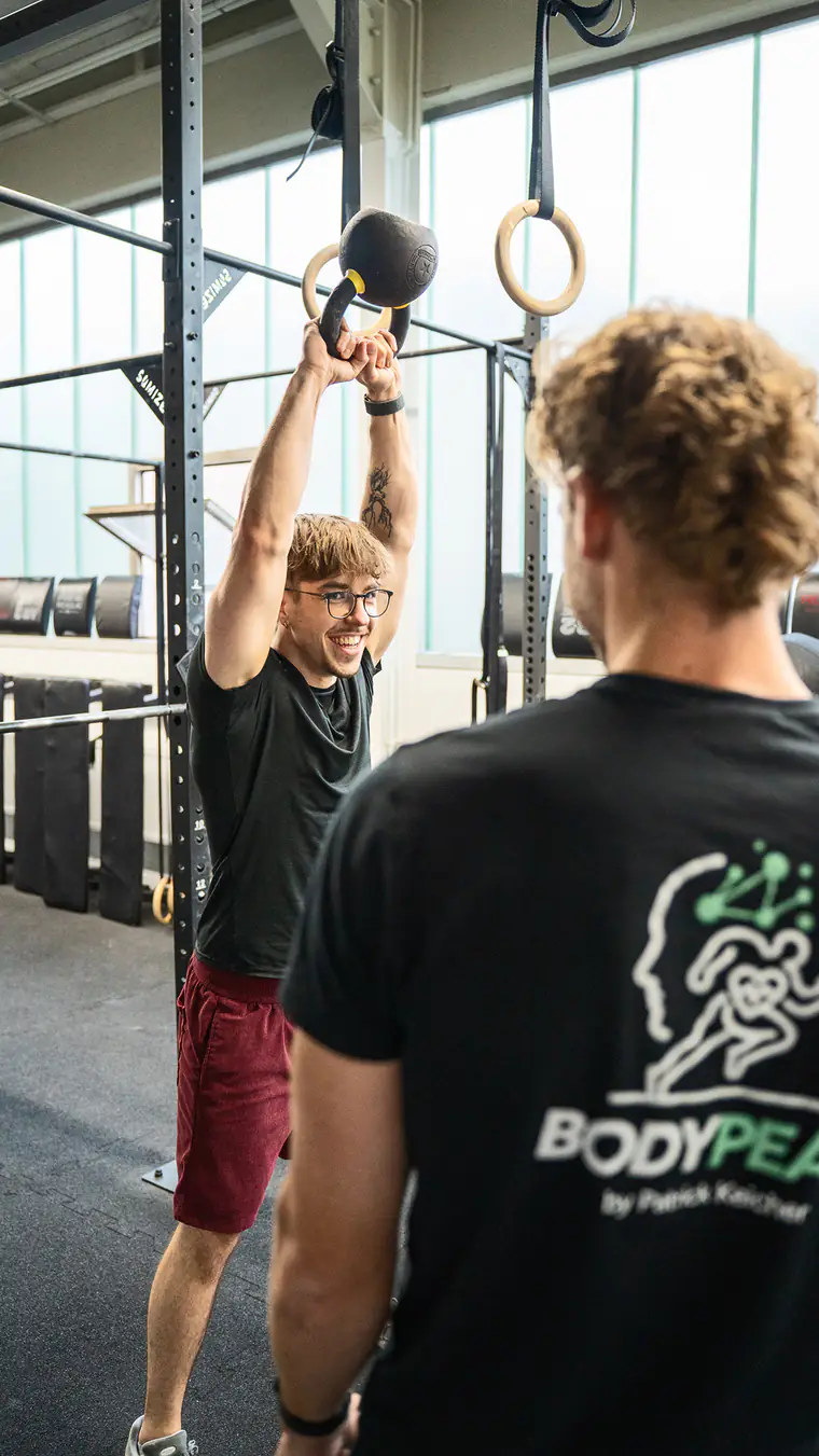 Ein Sportler drückt eine Kettlebell über den Kopf, Patrick Keicher steht daneben