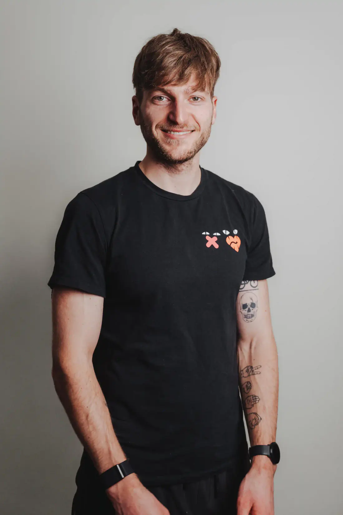 Patrick Keicher lächelt in die Kamera. Er trägt ein schwarzes T-Shirt mit farbigen Symbolen und steht vor hellem Hintergrund.