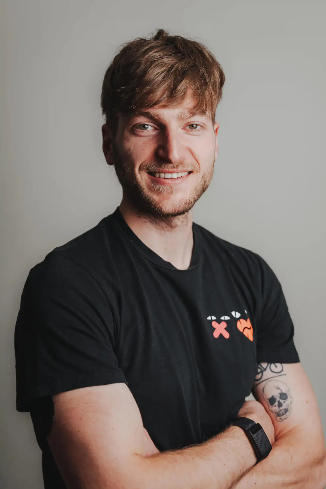 Patrick Keicher lächelt in die Kamera. Er trägt ein schwarzes T-Shirt mit farbigen Symbolen und steht vor hellem Hintergrund.