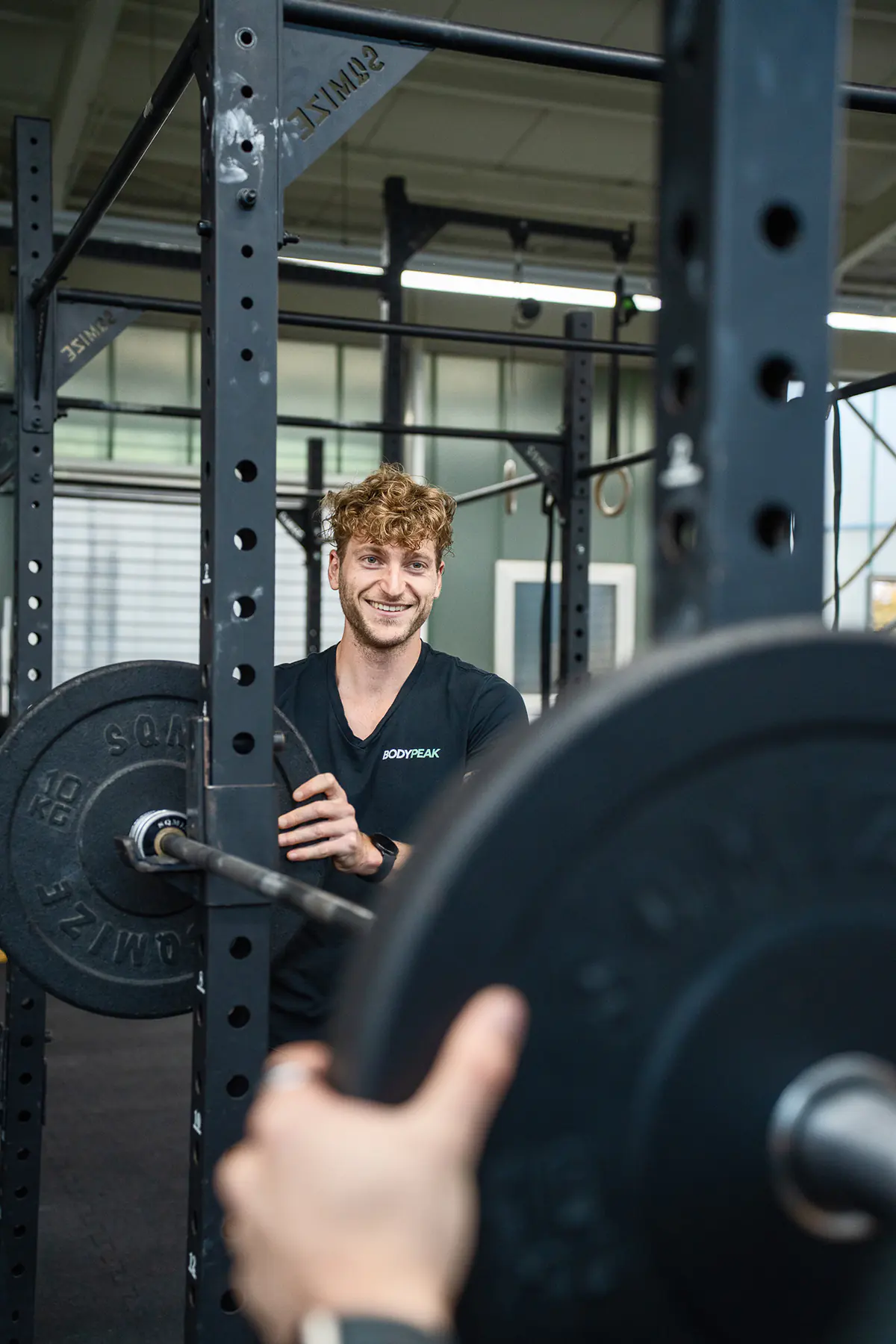 Patrick Keicher im schwarzen BodyPeak-Shirt lehnt lächelnd an einer Langhantel im Studio