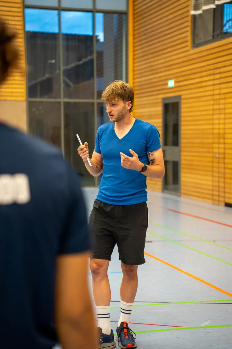 Patrick Keicher spricht in einer Sporthalle. Er trägt ein blaues T-Shirt, hält ein Lineal in der Hand und gestikuliert.